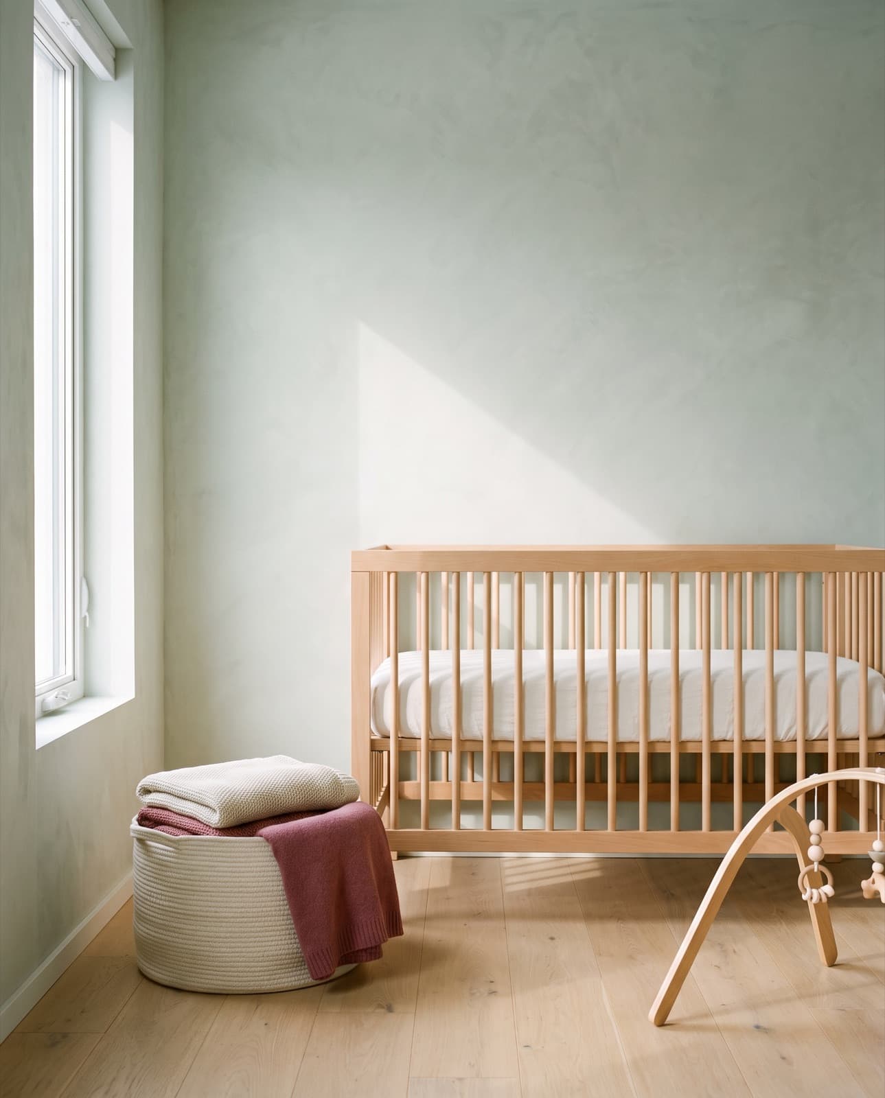 Chambre d'enfant au petit matin, lit à barreaux en hêtre clair, panier tressé et plaid rose sur un sol de chêne
